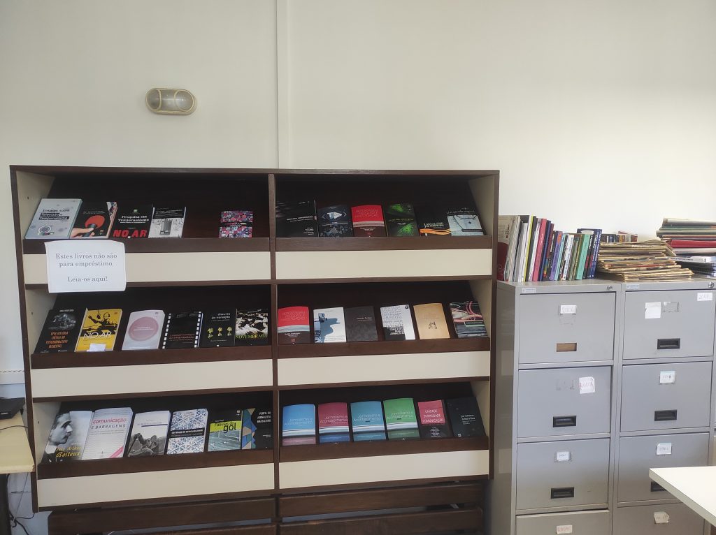 Hemeroteca do Curso de Jornalismo da UFSC. Na foto, há uma estante de livros ao lado de um armário que armazena alguns trabalhos realizados no curso.