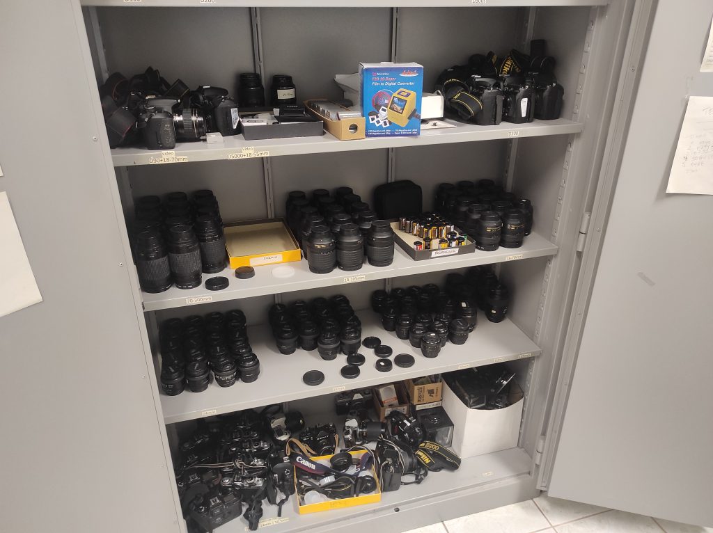 Armário aberto com equipamentos fotográficos: câmeras, rolos de filme, lentes, entre outros